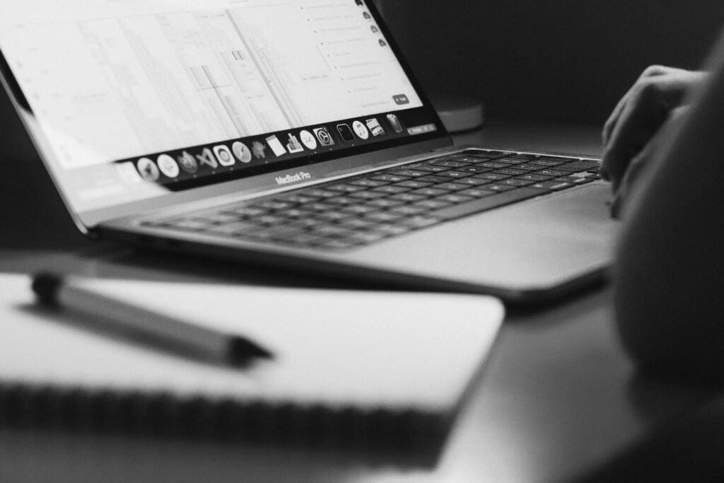 Black and white photo of a MacBook Pro on a desk, with a hand on the keyboard and a blurred notebook and pen in the foreground.