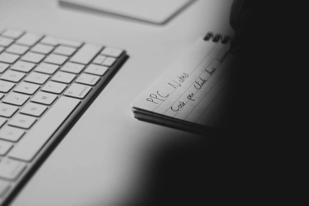 Black and white photo of a desk with a keyboard and a notepad titled ‘PPC Notes,’ showing someone writing about cost-per-click metrics